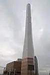 Ground-level view of a tall, white, angular chimney rising from a brown, striped building