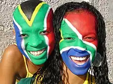 Dos personas con caras pintadas de la bandera de Sudáfrica. Llevan los colores de la bandera: verde, azul, rojo y negro, con líneas blancas y amarillas.