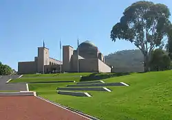 A view looking up a landscaped hill; steps lead to the entrance of a large cupola-topped building with three flag poles in front.