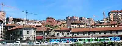 Vista exterior lateral, desde la orilla de la Ría. Se aprecia una unidad de tren de cercanías de Euskotren Trena estacionada en andén, junto a un tranvía en su ramal de servicio (hoy, prolongación hasta Bolueta)