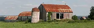 Un silo corto en el centro de la fotografía está ligeramente inclinado hacia la derecha, coronado por un techo rojo cónico. Tres graneros forman una V detrás del silo. A su derecha hay un granero grande, con techo rojo inclinado y puertas abiertas. Perpendicular a él hay dos graneros similares pero más pequeños en serie, visibles a la izquierda del silo. Graneros y silo en Newmarket, Ontario, Canadá. Estas estructuras fueron derribadas en marzo de 2009.