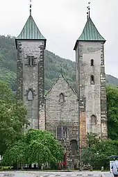 Una iglesia románica de piedra con dos torres y una entrada más baja de techo inclinado entre estas. Las dos torres tienen techos ligeramente curveados de bronce