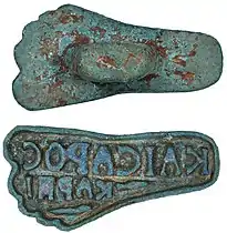 Sello de bronce; origen bizantino (s. IV-VI). En forma de pie e inscrito kaicapoc kap πo ('De César'). 8×4×2 cm.