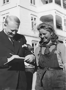 A middle-aged man in a dark coloured suit is writing on a pad of paper. Standing close to him and watching is a woman in her late twenties to earlier thirties. She is smiling, and wearing dark coloured dungarees, with a shirt underneath and a scarf tied around her neck.