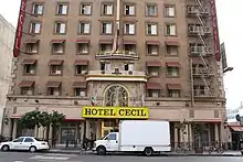 Los cinco pisos más bajos de un edificio de ladrillo tostado en una ciudad. Sus ventanas inferiores tienen toldos; hay una salida de incendios a la derecha. En la calle hay un gran refugio sobre la entrada con un cartel "Hotel Cecil" en negro sobre un fondo amarillo. En la calle enfrente están estacionados un pequeño camión y un carro, ambos blancos.