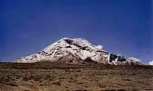 Una montaña nevada se halla ante un despejado cielo azul. El terreno circundante apenas posee algo de vegetación.