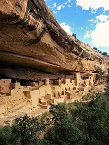 Imagen en color de unas grandes ruinas de arenisca, con árboles verdes abajo y cielo azul arriba