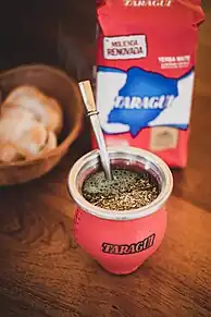 Mate de cuero rojo Taragui lleno de yerba mate Taragui y agua caliente para tomar un mate. Atras un paquete de yerba mate Taragui tradicional y unas tostadas en una cesta. Todo apoyado en una mesa de madera.