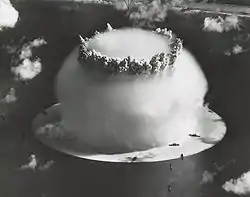 Aerial photo of nuclear explosion rising from lagoon. Hemispherical condensation cloud on the surface is 1 mile (1.6 km) in diameter. In comparison, Navy ships in the foreground look like bathtub toys.