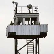 En la parte superior de una estructura de soporte tubular de acero con una escalera hay una pequeña cabina con ventanas a los lados, rodeadas de un stand en el balcón Qué dos hombres en uniforme, mirando al observador a través de binoculares; el tercer hombre está dentro de la cabina. Hay reflectores de techo plano de la torre, y la barandilla alrededor del borde exterior del techo.