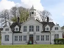 Una casa de madera ornamentada, simétrica, con dos pequeñas alas con columnas decoradas. El techo inclinado tiene frontones. Las ventantas se encuentran divididas y la puerta principal tiene columnas a ambos de sus lados.