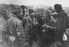 Un homme barbu en uniforme, coiffé d'une toque de fourrure, entouré d'autres hommes en uniforme.