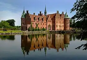 Un largo castillo de tres pisos tiene techos lanzados, dos torres circulares y una aguja, una larga entrada de frente al lago que refleja la imagen del castillo.