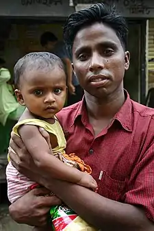 A young father dressed in a pink cotton shirt holds his child and gazes at the camera looking proud but tired. The little girl, wearing a sleeveless dress, sits on her father's arm and frowns directly at the camera.