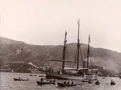  A three-masted ship, under steam power, moves across a stretch of water attended by several rowing boats. In the background is a line of hills, with buildings faintly visible at the water's edge.