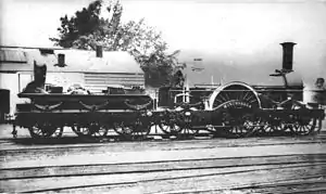 Una imagen en blanco y negro que muestra una locomotora de vapor hacia la derecha. El ténder abajo a la izquierda tiene seis ruedas; la máquina en sí tiene una rueda grande en el centro con dos ruedas delante y otra detrás.