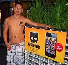 Un hombre musculoso con el torso desnudo posa al lado de un cartel publicitario de la aplicación Grindr.