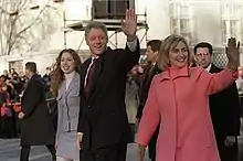 Same teenage girl, man and woman walk down a broad street in wintertime, as security personnel trail and a crowd looks on