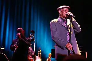 A man wearing a cap on his head, eye glasses, and a suit, holding on to a microphone stand with both hands. To the left of him is a man also holding on to a microphone stand. In between them are various musical instruments.