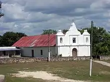 Vista sobre un campo abierto con parches de hierba gastada extendiéndose hacia una iglesia baja con una sencilla fachada blanca. La fachada tiene tres agujas cortas, la del medio más alta que las demás. La aguja central soporta una cruz. Las agujas más cortas cuentan con una abertura en forma de arco, una campana es visible en el arco de izquierda. La fachada cuenta con una puerta arqueada de madera; una ventana cuadrada está por encima de ella. La estructura principal de la iglesia tiene aproximadamente la mitad de la altura de la fachada y se extiende hacia atrás por el lado izquierda. La pared está lisa con excepción de una puerta más pequeña y una pequeña ventana. El techo es de metal corrugado oxidado. A la izquierda de la iglesia se encuentra un edificio bajo con una terraza sombreada. Dos postes soportan una línea eléctrica que pasa a través del campo a la iglesia. La base de cada poste está pintado de blanco. Una línea de árboles corre detrás de la iglesia.