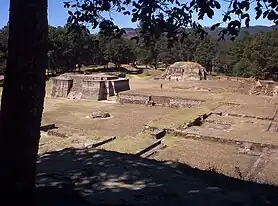 Vista de un número de ruinas bajas, bien mantenidos, que consisten en un laberinto de plataformas rectangulares basales superpuestas. Dos pequeñas estructuras piramidales dominan la vista, con un fondo de bosque de pinos.