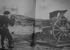 Fotografía en blanco y negro de Churchill uniformado mirando a través del cañón de un gran arma de artillería con una barricada de piedra visible detrás