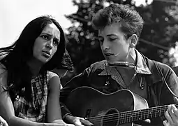 Outdoors,Joan Báez is sitting next to Bob Dylan who is playing an acoustic guitar, ca 1960s.