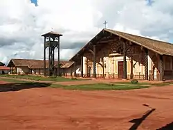 A church and bell-tower in frontal view. The whitish facade is decorated with motives painted in orange. A cross is positioned at the top of the roof.