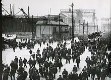 Trabajadores de Harland and Wolff a la salida del astillero, hacia abril de 1911.  A la izquierda, se observa el SS Nomadic, transbordador del Olympic y del Titanic, durante su acondicionamiento. Al fondo, se observa el pórtico Arrol, con la proa del Titanic perfectamente visible.