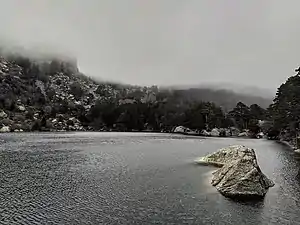 El agua contiene decenas de ondas provocadas por el viento. Al fondo de la foto se ve la cima de la montaña tapada por la niebla, que desciende hasta casi tocar el agua. La ladera de la montaña está manchada por trozos de nieve.