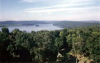 Una vista a través de la selva hacia un lago rodeado de colinas boscosas bajas. Dos pequeñas islas se encuentran cerca de la orilla opuesta. Hay un pequeño claro en el primer plano inmediato. En el lado izquierdo de la orilla opuesta se encuentra una amplia área herbosa frente a las colinas boscosas.