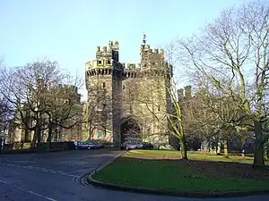 View down a short tree-lined drive leading to the main entrance of a castle, in front of which there are parked cars. The sky is blue and cloudless, but the shadows are long and the trees are not in leaf.