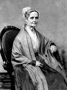 A monochrome photograph portrait of an elderly woman seated in a chair, wearing a heavy shawl over a white blouse, a simple lace bonnet covering hair held up on the back of the head, the woman turned somewhat to the right