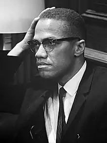 An African American man in his forties, wearing glasses and a suit and tie, sitting and looking to the right, with his hand resting on his right temple.