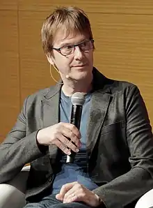 Fotografía de Mark Cerny, un hombre estadounidense de mediana edad que sostiene un micrófono y lleva gafas, camiseta azul y abrigo negro.