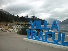 Fotografía que muestra el nombre de la localidad en letras grandes. Detrás se distingue el Lago Traful con algo de oleaje. A la izquierda de la imagen se encuentra el muelle (en menor tamaño puesto que se sitúa a varios metros de distancia), y en el fondo se aprecian los árboles de pino (entre otras especies) y las montañas en un día grisáceo, completamente nublado.