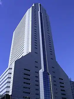 Ground-level view of a light blue high-rise; one flat facade is bisected by a column of glass, while the adjacent facades angle in at several points until they meet at a point at the top