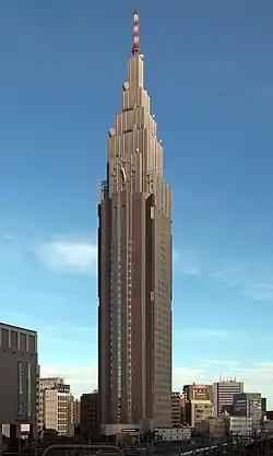 Ground-level view of two of a building's faces, both with many different facets, set in front of a dark blue sky; as the building rises, it terraces to a point and a white and an orange antenna rises from the top. A clock is located on one side of the building.