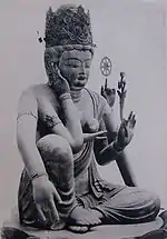 Three-quarter view of a seated statue with six arms. The left leg is bend, touching the ground along its whole length. The right leg is also bend, but with the knee up. The right foot is above the left foot. One of the right hands is touching the right ear, another is lifting a jewel-shaped object in front of the breast. One of the left hands carries a flower, another a small wheel-shaped object.