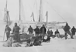 Un grupo de hombres posan sobre el hielo con perros y trineos, con la figura de un barco visible en el fondo