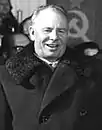 A man wearing a long coat, smiling, in front of a group of people and the Soviet flag