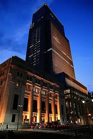 Ground-level view of a rectangular, glass high-rise; adjoining the high-rise is a stone building featuring columns