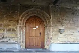 La puerta principal por la que se accede a la Ermita es de medio punto, con tres arquivoltas estriadas con capiteles lisos y flanqueada por robustas columnas también estriadas y rematadas con capiteles idénticos. A la izquierda de la puerta se puede ver la aguabenditera junto con dos bancos corridos de piedra adosados a la pared, en ambos lados.