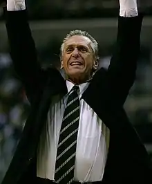 Pat Riley, wearing a black suit with a dark green and silver tie, is throwing his hands in the air as he celebrates a good play.