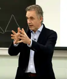 Jordan Peterson en un traje negro con camisa azul haciendo un gesto con las manos casi cerrándolas en un salón de clases al frente de un tablero de tiza