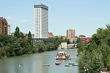 El río Pisuerga a su paso por Valladolid, con el Puente Mayor al fondo. En el medio se encuentra el barco. A su izquierda el Edificio Duque de Lerma y en frente se sitúa la Playa de las Moreras.