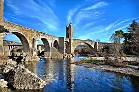 Puente de Besalú, de forma angular con siete arcos desiguales soportados por pilastras sobre la roca viva, con tajamares. Original románico anterior a 1071, ha sido reconstruido muchas veces.