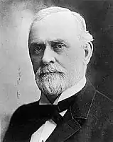 headshot image of a Caucasian male with white hair and a goatie who is wearing a tuxedo with a black tie.