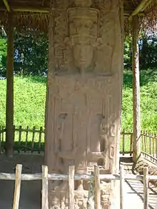 Una estela con la prominente escultura de un rey rodeado de decoración elaborada. El monumento está cubierto por un techo de paja sostenido por postes de madera.