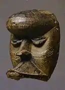 Mascarilla zooantropomórfica con frente fuertemente redondeada y ojos rasgados que sobresalen; incisiones curvilíneas superpuestas que se ramifican a la altura de la nariz. Mandíbulas de animales proyectadas hacia adelante (falta la parte inferior móvil). Agujeros para sujetar la máscara al disfraz a lo largo del contorno. Las máscaras que mezclan elementos humanos y animales son manifestaciones de los espíritus del bosque.